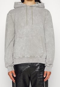 Felpa grigia chiara con una finitura testurizzata, cappuccio con cordino e tasca frontale. Indossata con pantaloni neri lucidi in pelle sintetica a stampa coccodrillo.