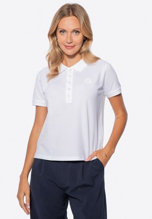 Weißes Poloshirt mit Kragen, ausgestattet mit einer Knopfleiste und einem runden Logo-Detail. Getragen mit marineblauen Hosen, glatte Textur.