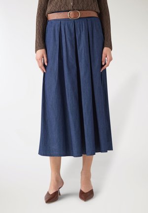 Donna con gonna midi plissettata blu scuro, maglione marrone lavorato a trama, cintura color cuoio e scarpe con tacco marroni a punta.