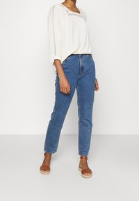 Helle cremefarbene Bluse mit quadratischem Ausschnitt und elastischen Ärmeln; kombiniert mit hoch taillierten blauen Jeans und braunen Sandalen mit Riemchen.