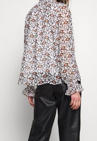Transparante blouse met een zwart, wit en oranje abstract patroon, met ruches aan de manchetten en een losse pasvorm, gecombineerd met zwarte leren broek.