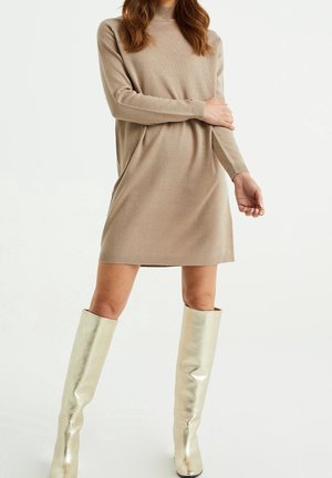 Femme en robe pull beige à manches longues et bottes crème brillantes montant jusqu'aux genoux, debout devant un fond blanc uni.