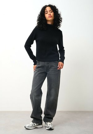 Zwarte coltrui met ribbeltextuur, gecombineerd met hoogwaisted donkergrijze spijkerbroek. Model draagt zilveren sneakers met zwarte accenten.