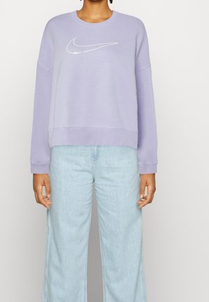 Personne portant un sweat-shirt violet clair avec un logo Nike Swoosh blanc à pois et un jean bleu clair à jambes larges sur un fond uni.