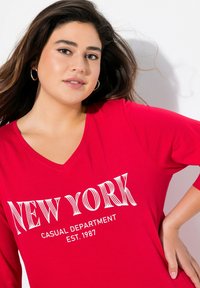 Rotes Baumwoll-T-Shirt mit V-Ausschnitt, mit den Worten "NEW YORK" in fettem weißen Text und dem Satz "CASUAL DEPARTMENT EST. 1987" darunter.