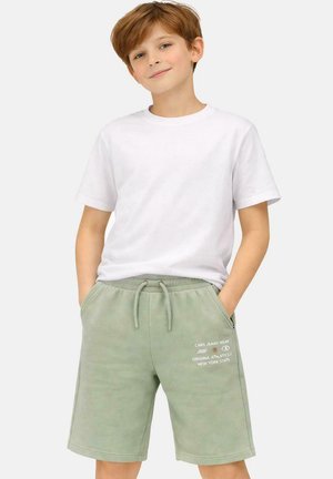 Jongen met rood haar, gekleed in een wit T-shirt met korte mouwen en lichtgroene short met trekkoord, met handen in de zakken.