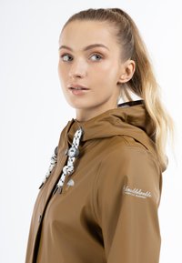 Giacca impermeabile marrone con cappuccio, dotata di lacci bianchi e neri, dettagli in argento e un logo sulla manica. Tessuto liscio.