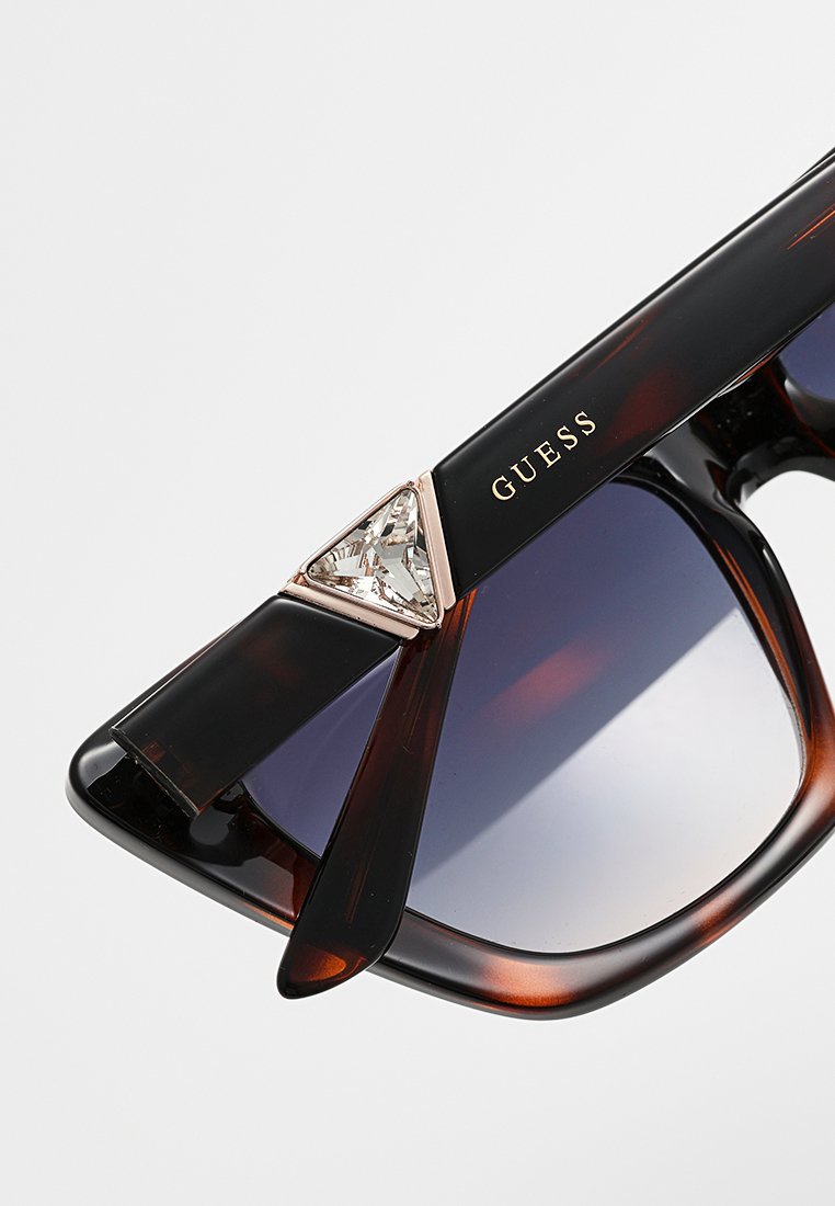 Lunettes de soleil avec une monture noire et écaille de tortue, des verres dégradés, et un accent en cristal triangulaire sur la branche. Logo "GUESS" sur le côté.