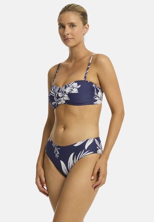Kobieta ubrana w granatowy bikini z białym kwiatowym wzorem, stojąca na tle jednolitego jasnego tła, lekko zwrócona na prawo.