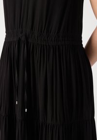Robe noire avec taille froncée et cordon fin à nouer dans le dos, montrant une partie du bras et du torse d’une personne.
