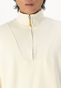 Pullover crema con mezza cerniera, collo alto, trama a coste, vestibilità squadrata e cerniera visibile con cursore giallo.