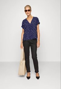 Mulher vestindo blusa estampada azul-marinho, calças pretas, saltos pretos, óculos de sol, segurando uma grande bolsa bege, posando contra um fundo liso.