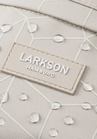 Borsa strutturata beige con superficie testurizzata caratterizzata da punti rialzati lucidi, linee geometriche bianche e una patch rettangolare con logo in rilievo.