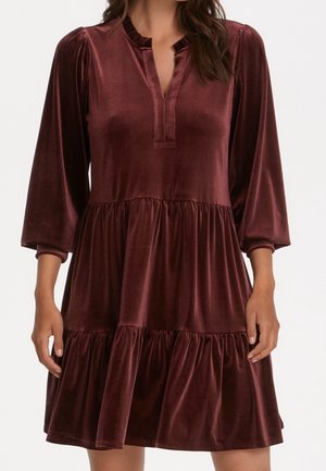 Femme portant une robe longue à manches longues en velours rouge foncé à étages, avec un décolleté fendu et un col à volants, debout les bras détendus.