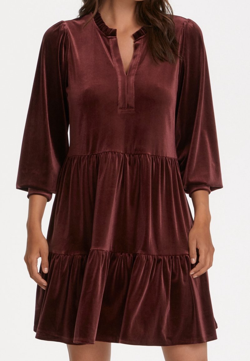 Femme portant une robe longue à manches longues en velours rouge foncé à étages, avec un décolleté fendu et un col à volants, debout les bras détendus.