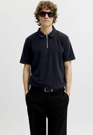 Jeune homme aux cheveux bouclés portant des lunettes de soleil noires, une chemise sombre à manches courtes avec col zippé et un pantalon noir, les mains dans les poches.
