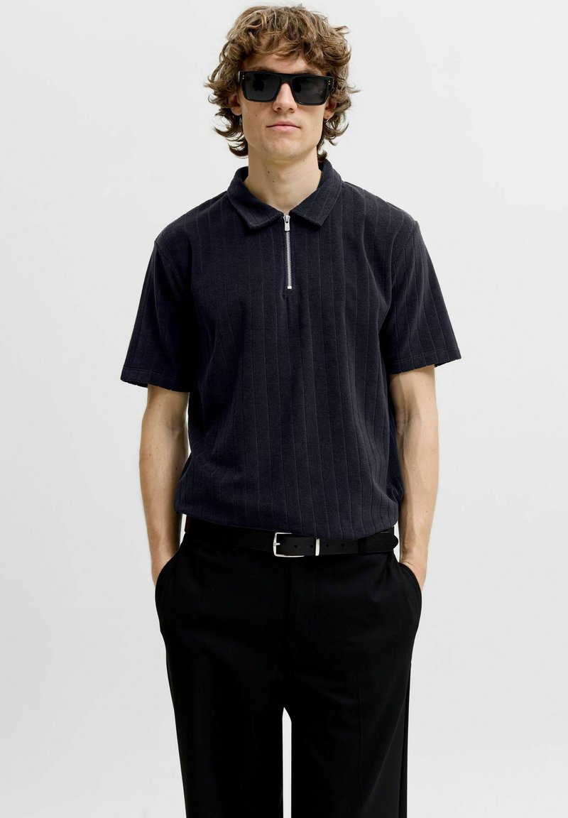 Jeune homme aux cheveux bouclés portant des lunettes de soleil noires, une chemise sombre à manches courtes avec col zippé et un pantalon noir, les mains dans les poches.