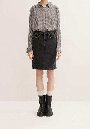 Personne portant une chemise à carreaux à manches longues, une jupe en denim noire taille haute, des chaussettes crème et des bottines noires, debout devant un fond uni.