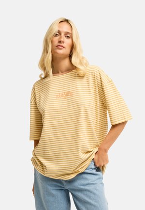 Blonde Frau trägt ein lockeres gelb-weiß gestreiftes T-Shirt und hellblaue Jeans, steht mit einer Hand in der Tasche.