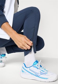 Jūriniai sportiniai džinsai su kulkšnių užtrauktukais, derinami su baltais ir mėlynais sportiniais batais bei baltais audiniais kojinėmis. Lygi medžiagos tekstūra.