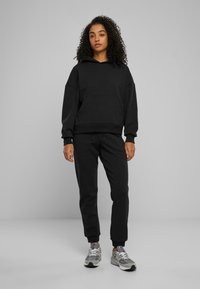 Felpa nera con cappuccio, grande tasca frontale e polsini a coste abbinata a pantaloni jogger coordinati. Modello indossa scarpe da ginnastica grigie.