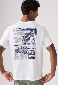 Λευκό βαμβακερό μπλουζάκι με μπλε γραφικό εκτύπωση στην πλάτη, απεικονίζοντας μια δυτική σκηνή με κείμενο και τη σήμανση "Levi's".