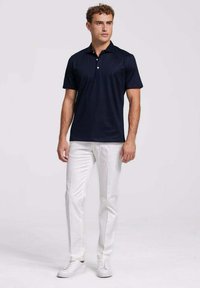 Marineblauwe polo met korte mouwen en een kraag met drie knopen, gecombineerd met witte broek en witte sneakers, staande tegen een effen achtergrond.