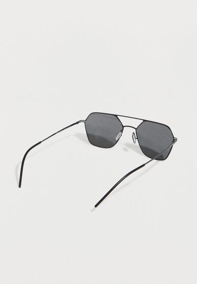 Μαύρα γυαλιά ηλίου με μεταλλικό σκελετό και εξαγωνικούς φακούς με σκούρο φίλτρο, και λεπτούς βραχίονες που ακουμπούν σε λευκή επιφάνεια.