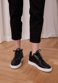 Černé kožené tenisky se zlatými očky a detaily, bílé gumové podešve a elegantním nízkým designem. Texturovaný svrchní detail.