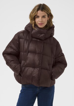 Brun polstret jakke med høj krave, der har et quiltet design, justerbare snore og sidelommer. Båret med blå jeans.