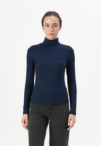 Marineblauwe geribde coltrui met lange mouwen en een aansluitend ontwerp, met een klein logoaccent op de borst. In combinatie met donkergrijze broek.