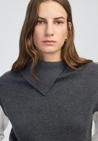 Grijze trui met een gestructureerde brei en een driehoekige kraag, gelaagd over een wit shirt. Glad materiaal en een aansluitende silhouet.