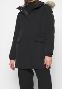 Parka nera con cappuccio foderato in pelliccia, dotata di chiusure a zip e a scatto, multiple tasche frontali e una texture trapuntata.