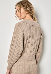 Beżowy sweter z dzianiny w warkocz z szerokim kołnierzem, ściągaczami przy rękawach i w pasie. Charakteryzuje się teksturowanym wzorem i luźnym krojem.