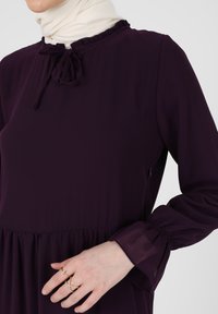 Robe violette à manches longues avec un col à volants et un nœud. Tissu léger ; manches évasées avec un détail froncé. Design minimaliste.