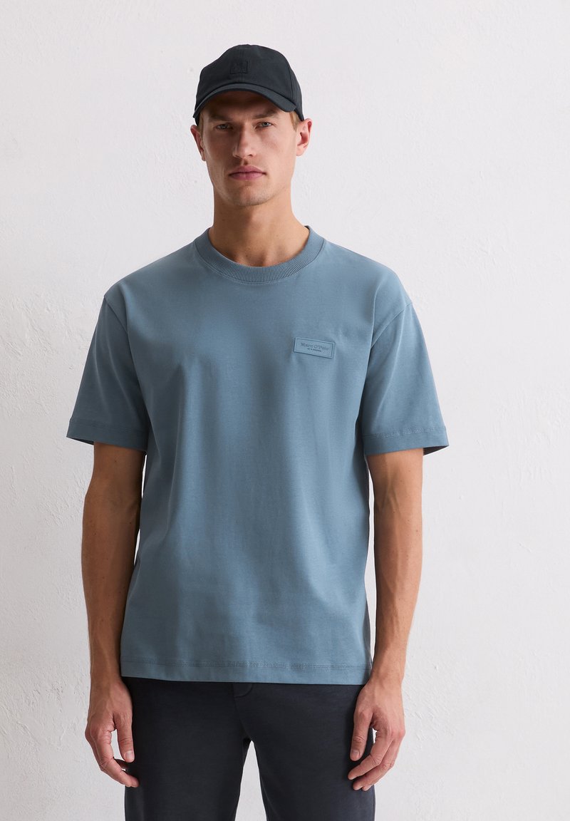 T-shirt en coton bleu clair avec un col rond, des manches courtes, et un petit patch logo rectangulaire sur la poitrine. Texture lisse.