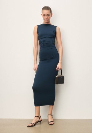 Nő áll egy sima fal előtt, viselve testhezálló, ujjatlan tengerészkék midi ruhát, fekete pántos magas sarkú cipőt, és tart egy kis fekete kézitáskát.