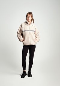 Giacca in pile beige con chiusura a zip, caratterizzata da una fascia con logo nero sul petto. Abbinata a leggings neri e sneaker nere.