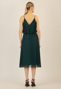 Grünes, plissiertes Kleid mit tiefem V-Ausschnitt hinten, verstellbaren dünnen Trägern und Mid-Calf-Länge. Durchsichtiger Überzug am Saum und taillierter Schnitt.