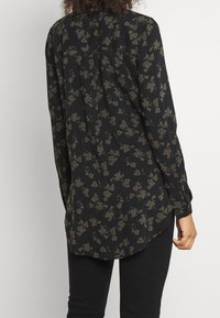Blusa negra de flores con mangas largas, que presenta un patrón floral verde, dobladillo curvado y puños con botones. Fabricada en tejido suave.