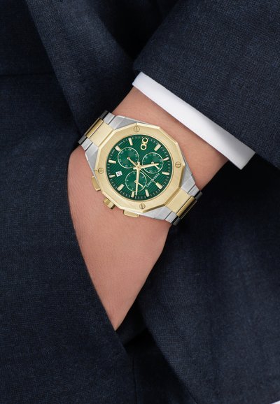 Zöld számlapos óra ezüst-arany színű, nyolcszögletű tokkal, ezüst-arany karkötővel és három segédszámlappal. Egy sötét ruhát viselő csuklón látható.