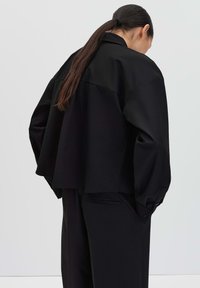 Vrouw met lang haar, gekleed in een losse zwarte jas en bijpassende wijde broek, met haar rug naar de camera gekeerd tegen een vlakke lichte achtergrond.