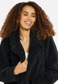 Cappotto nero peloso con colletto a revers, chiusura anteriore con bottoni e texture morbida. Il modello mostra il cappotto parzialmente aperto, rivelando una maglietta bianca sotto.