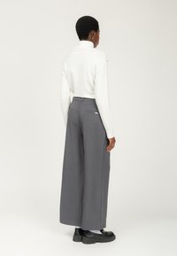 Persona con cabello corto vestida con un suéter blanco de cuello alto, pantalones anchos grises, calcetines blancos y mocasines negros gruesos de pie de espaldas.