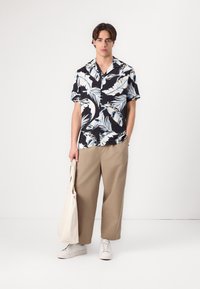 Camicia nera a maniche corte con motivo di foglie di palma bianche e azzurro chiaro; pantaloni larghi khaki; sneakers bianche; tiene una borsa tote color crema.