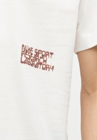 Λευκό κοντομάνικο μπλουζάκι με κόκκινο κείμενο που αναγράφει "NIKE SPORT RESEARCH LABORATORY" στο πάνω αριστερό μέρος του στήθους, φορεμένο από ένα άτομο με ορατό χέρι.