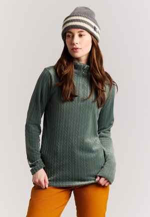 Junge Frau, die eine graue Strickmütze, einen grünen Pullover mit Reißverschluss und strukturiertem Muster sowie senfgelbe Hose trägt und vor einem einfarbigen Hintergrund steht.