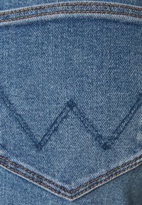 La tela de mezclilla con un tono azul presenta un distintivo patrón de costura triangular en el bolsillo trasero, acentuado por hilo marrón contrastante.