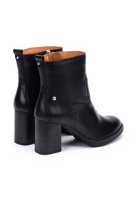 Botas de cuero negro hasta el tobillo con tacón apilado, que cuentan con costuras decorativas y detalles metálicos. Textura suave con un diseño minimalista.