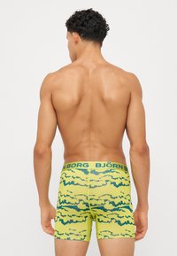 Dzelteni bokseršorti ar zaļiem mākoņu rakstiem, kuriem ir plata elastīga josta ar "BJÖRN BORG" zīmolu, izgatavoti no elastīga auduma.